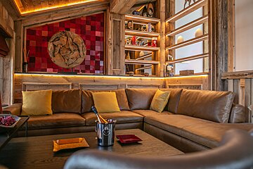A cozy, rustic living room with a large brown leather sectional, wooden walls, backlit shelves, and a red checkered art piece. Champagne awaits.