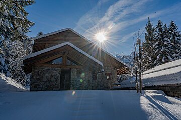 Snowy stone and wood chalet under a bright winter sun. Pine trees and mountains frame the cozy scene.