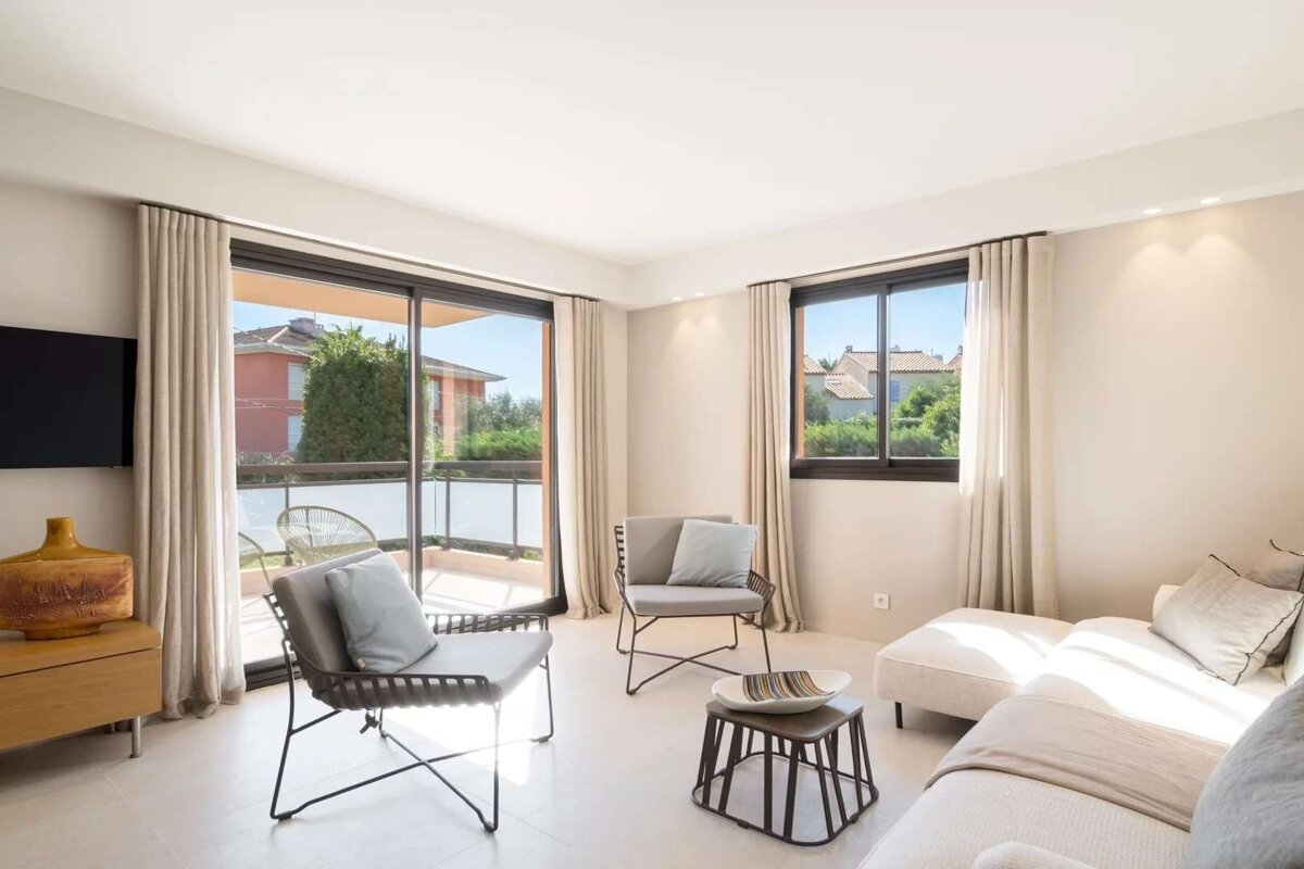 A bright, modern living room with light decor, large windows and sliding doors opening to a balcony with views of houses and trees.