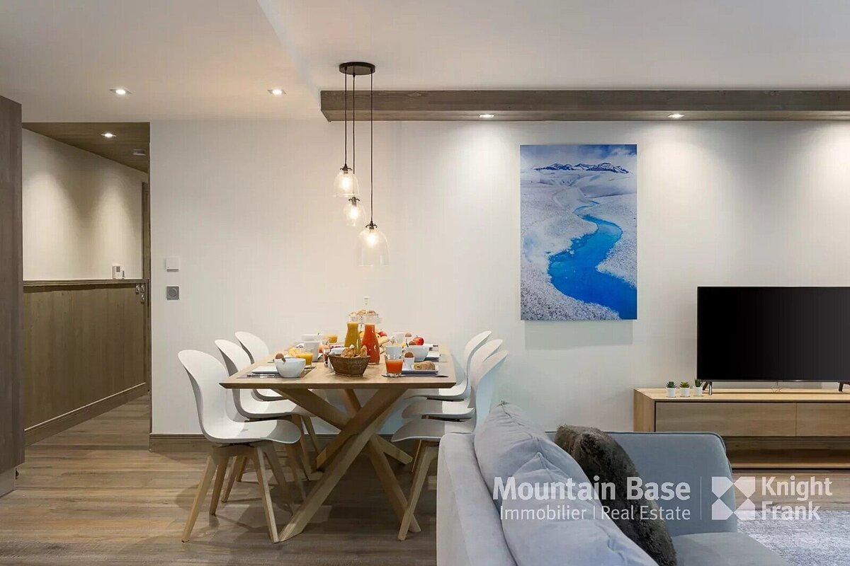 A modern dining area with a wooden table set for breakfast, white chairs, and pendant lights. A large blue river artwork hangs above a TV and sofa, all on wooden floors.