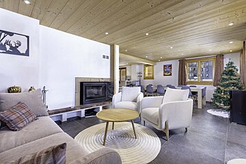 A spacious, cozy chalet-style living and dining room with a fireplace, modern furniture, wooden ceiling, and a decorated Christmas tree.