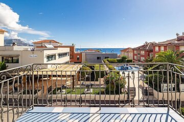 Balcony view of coastal buildings, a playground, and the sea with boats under a clear blue sky, with distant mountains.
