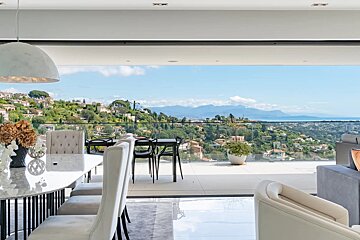 A modern room with dining furniture and a sofa, featuring a stunning panoramic view of green hills, distant houses, and the sea under a blue sky.