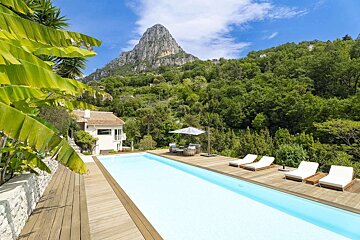 A luxury infinity pool with a wooden deck, white house, and tropical foliage, set against a stunning backdrop of lush green mountains and a peak.