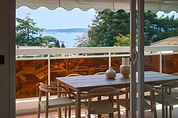 A balcony dining area with a wooden table and chairs, offering a scenic view of the sea and lush green trees.