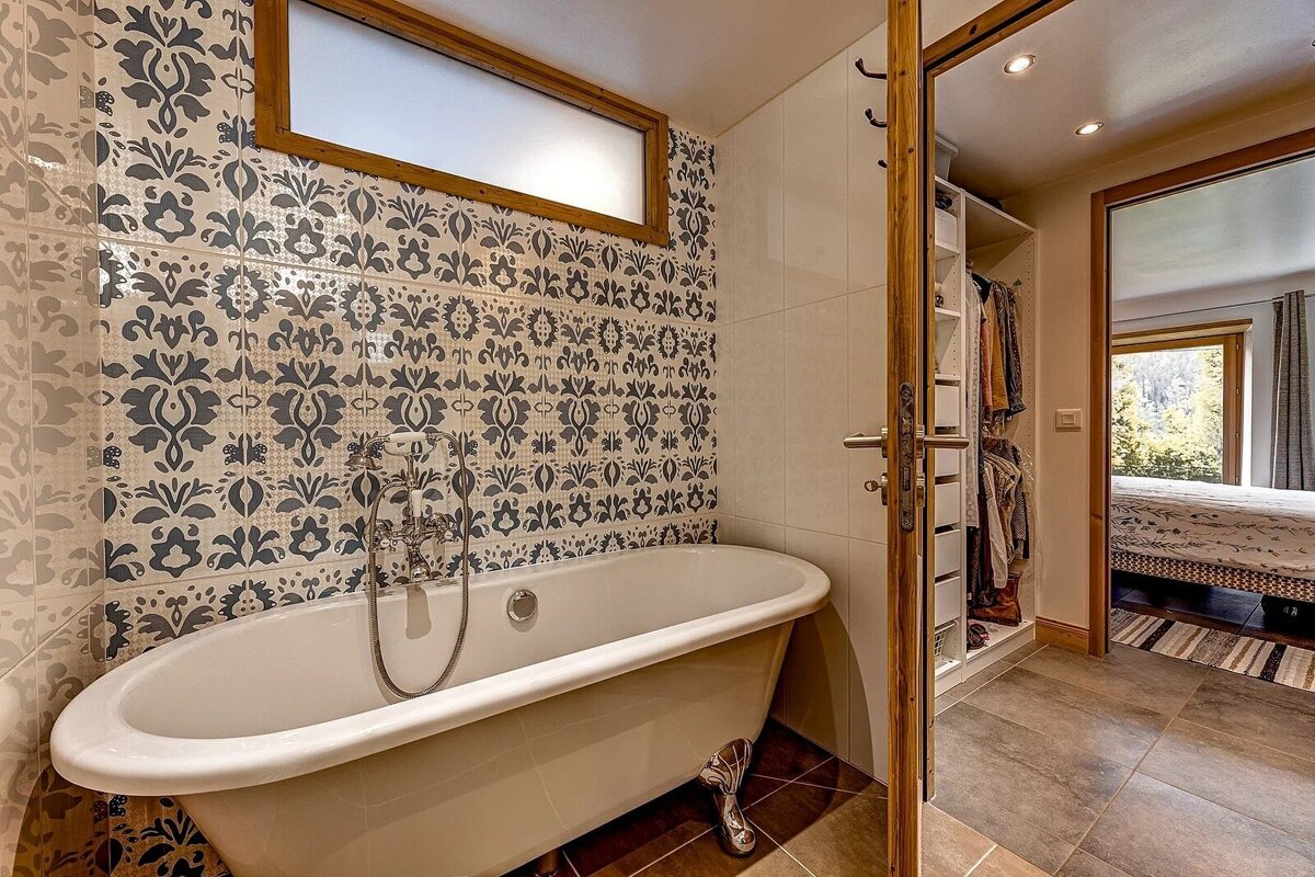 A white clawfoot bathtub against a patterned tile wall, with an open doorway revealing a closet and a bedroom beyond.