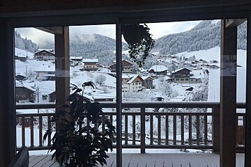View from a window of a snow-covered mountain village with chalets, surrounded by snowy trees and mountains. A balcony railing and plants are in the foreground.