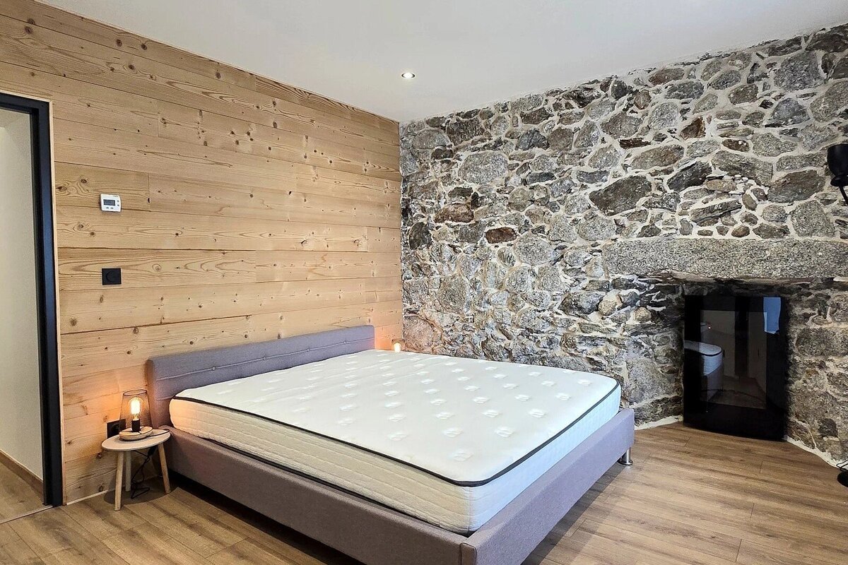 Rustic bedroom featuring a wood-paneled wall, a stone accent wall with a fireplace, and a grey bed.