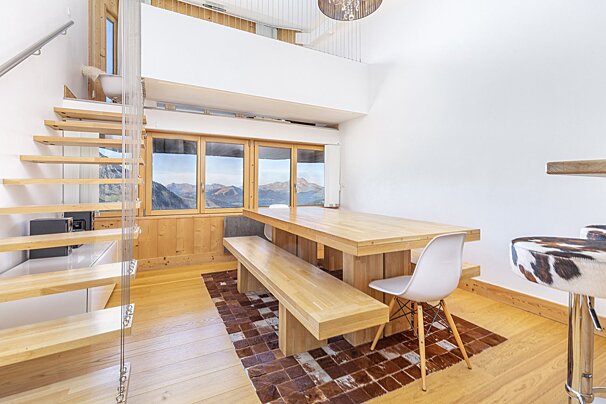 Modern chalet interior with a wooden dining table, floating stairs, and a mountain view through windows.