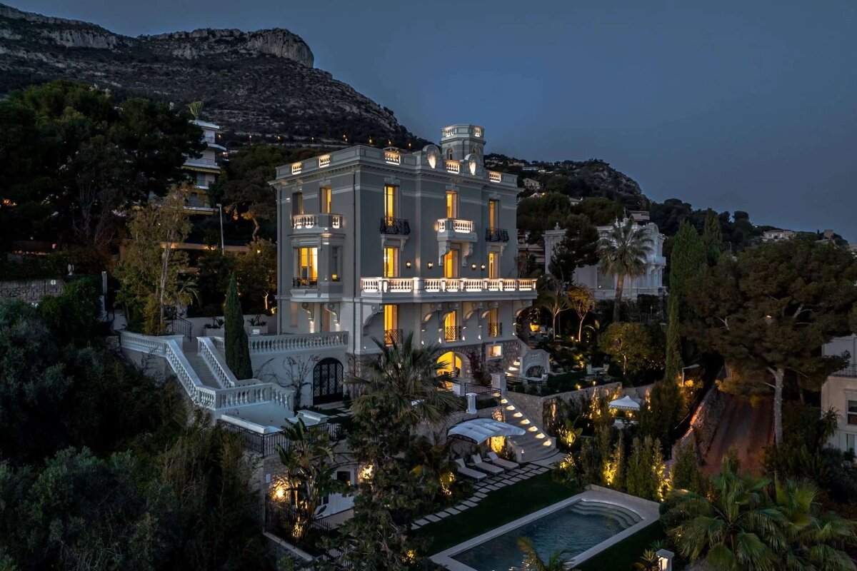 A stunning multi-story villa glows at dusk on a mountainside, with lit windows, balconies, a pool, and tiered gardens creating an elegant scene.