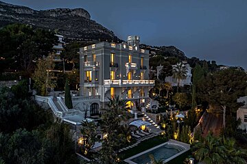 A stunning multi-story villa glows at dusk on a mountainside, with lit windows, balconies, a pool, and tiered gardens creating an elegant scene.