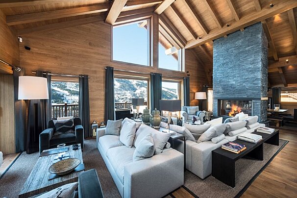 Luxurious wood-paneled chalet living room with a stone fireplace, modern sofas, and expansive windows overlooking snowy mountains.