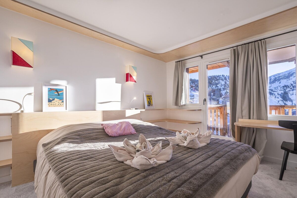 A bright, modern bedroom with a fluffy bed, geometric wall lights, and a balcony offering a panoramic view of snowy mountains and a ski resort.