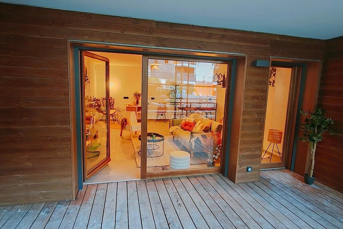 A wooden deck connects to a wood-clad building through large glass doors, revealing a warm and inviting interior living space.