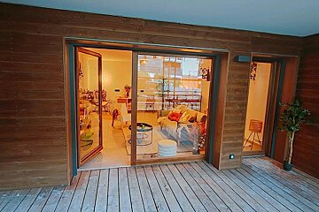 A wooden deck connects to a wood-clad building through large glass doors, revealing a warm and inviting interior living space.