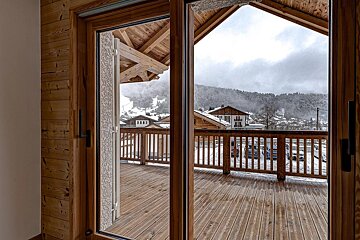 View through a wooden glass door of a balcony overlooking a snowy mountain village under a cloudy winter sky.