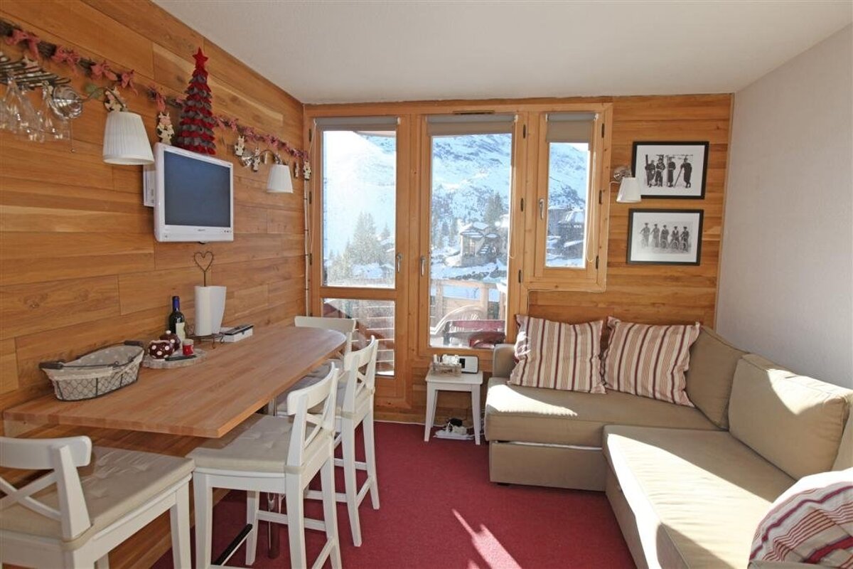 Cozy wood-paneled chalet room with a bar-height table, sofa, and TV. Large windows offer snowy mountain views. Festive Christmas decor.
