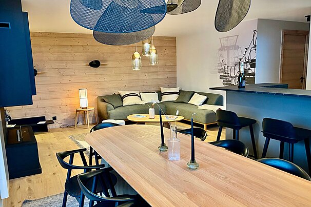 A modern open-plan living and dining area with wooden walls, a grey sectional sofa, light wood dining table, and bar. Blue mesh chandeliers and a ski lift mural add character.
