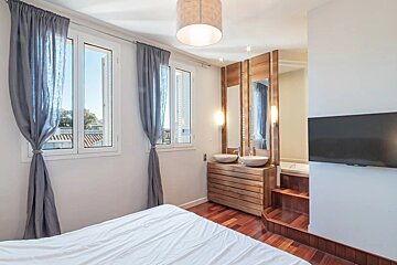 Modern bedroom with white bed, gray curtains, wood vanity with double sinks & mirrors, elevated tub, and wall-mounted TV. Hardwood floor.