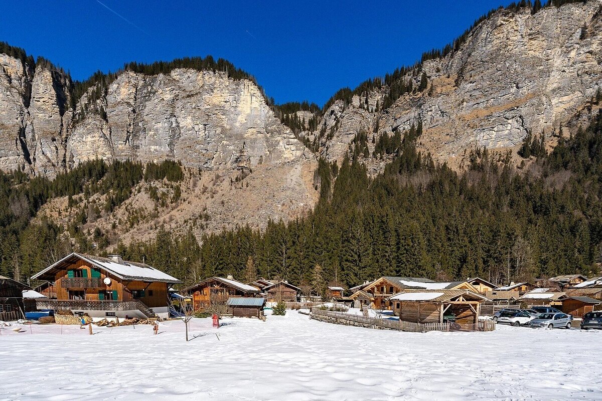 Snow-covered village of rustic wooden chalets nestled at the base of towering, tree-lined mountains under a vivid blue sky.