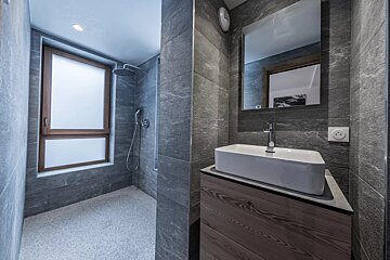 Sleek, modern bathroom with dark gray tiles, featuring a walk-in shower with a window and a vanity with a rectangular sink and mirror.