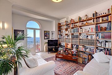 A bright living room with white sofas, extensive bookshelves, and an arched glass door leading to a balcony overlooking a stunning sea view.