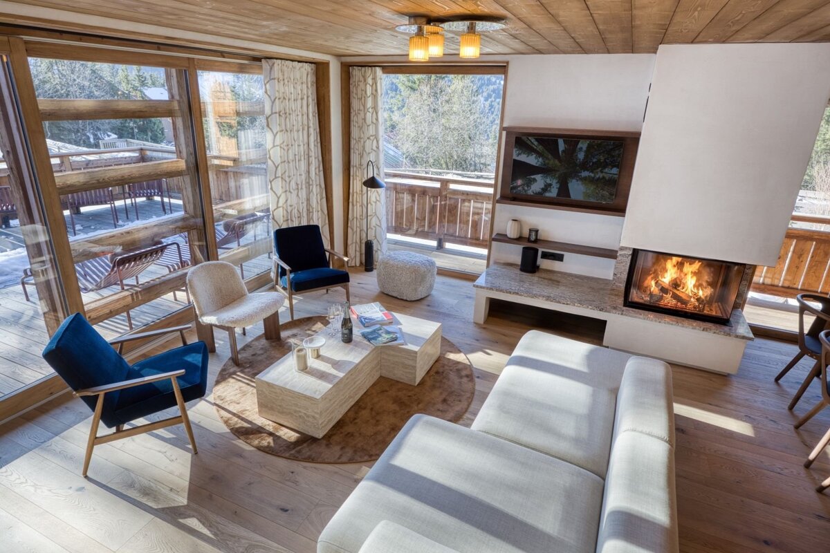 Modern living room with a lit fireplace, comfy seating, wood accents, and large windows revealing a snowy outdoor balcony and forest.