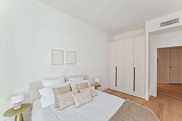 A bright, minimalist bedroom featuring a bed with neutral-toned pillows, white walls, built-in wardrobes, and hardwood flooring.