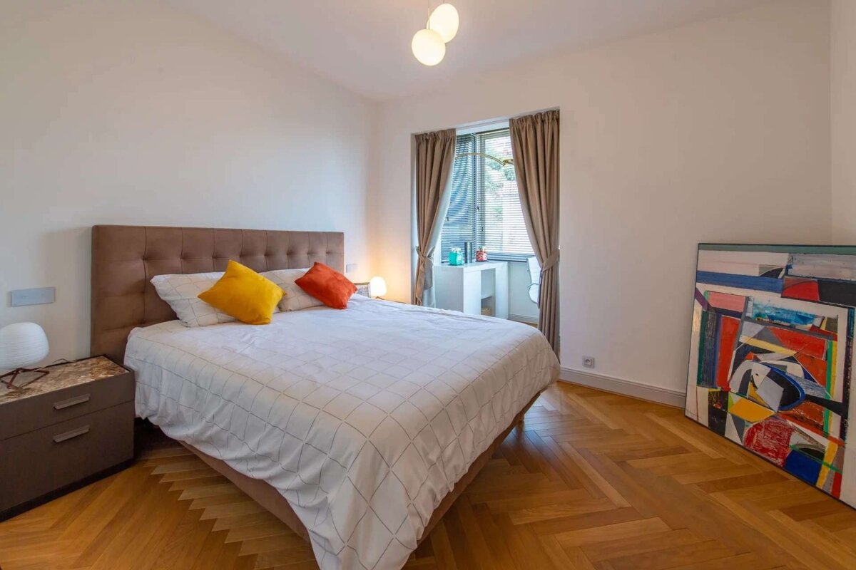 A modern bedroom features a bed with colorful pillows, a wooden herringbone floor, and a window with blinds and curtains. A vibrant abstract painting leans against the wall.