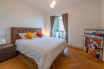 A modern bedroom features a bed with colorful pillows, a wooden herringbone floor, and a window with blinds and curtains. A vibrant abstract painting leans against the wall.