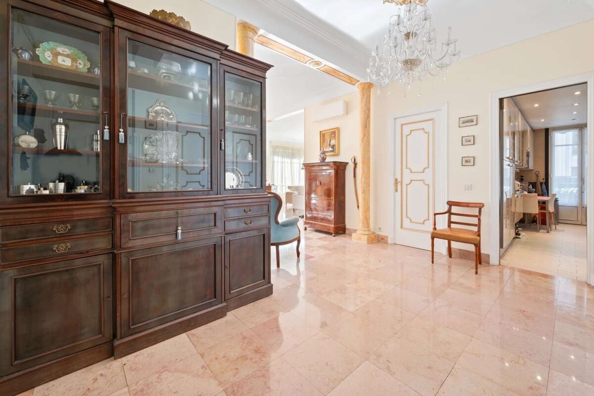 An ornate interior features a large wooden display hutch, marble floors, a crystal chandelier, gilded doors, and classical columns, leading to other rooms.
