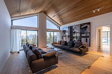 Spacious, modern living room with dark sofas, a wooden ceiling, and floor-to-ceiling windows offering panoramic views of snowy mountains and pine trees.