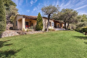 A warm-toned house with a red-tiled roof, surrounded by a lush green lawn and mature trees under a bright blue sky.