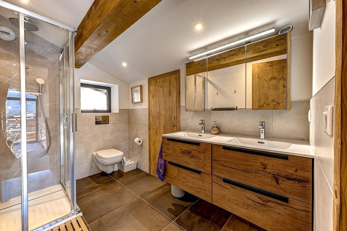 Modern rustic bathroom featuring a glass shower, double-sink wooden vanity with mirrored cabinet, and toilet. Exposed wood beams and tile accents complete the look.