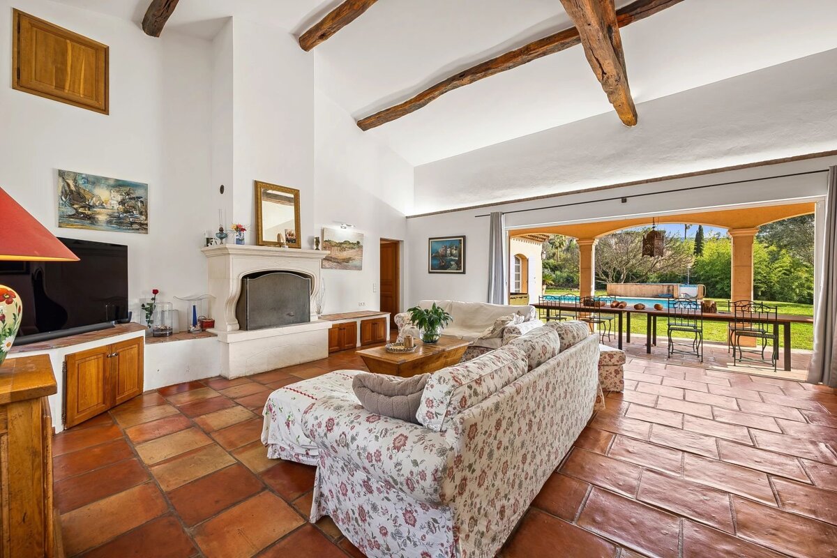 Spacious living room with terracotta tiles, exposed beams, a fireplace, and a large opening onto a terrace with a pool.