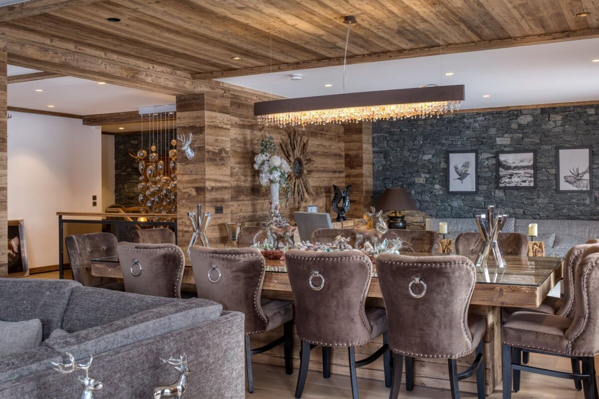 Chic lodge dining room with a long wooden table, plush velvet chairs, wood and stone walls, and elegant deer-themed decor under a sparkling chandelier.