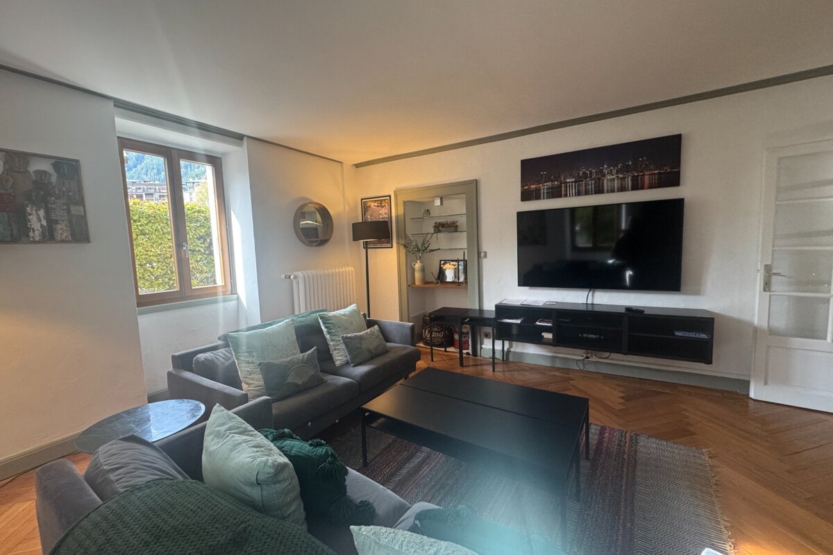 A modern living room with gray sofas, green pillows, a large wall-mounted TV, black furniture, and a window revealing mountains & greenery.
