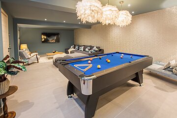 A modern game room featuring a blue pool table, unique feathery pendant lights, and a cozy lounge area with a sofa and TV.