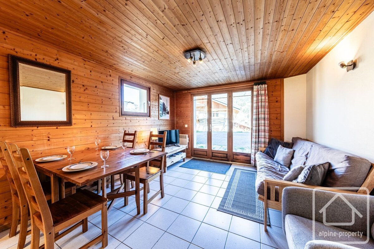 A cozy, wood-paneled room with a rustic dining table set for six, a futon sofa, and glass doors opening outside. Features a white tiled floor.