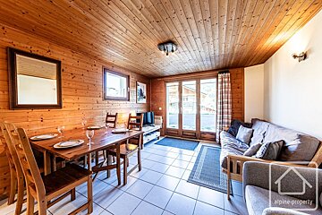 A cozy, wood-paneled room with a rustic dining table set for six, a futon sofa, and glass doors opening outside. Features a white tiled floor.