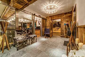 Rustic chalet lobby featuring grey stone floors, cowhide chairs, wooden walls, a round light fixture, and an open door showing snow outside.