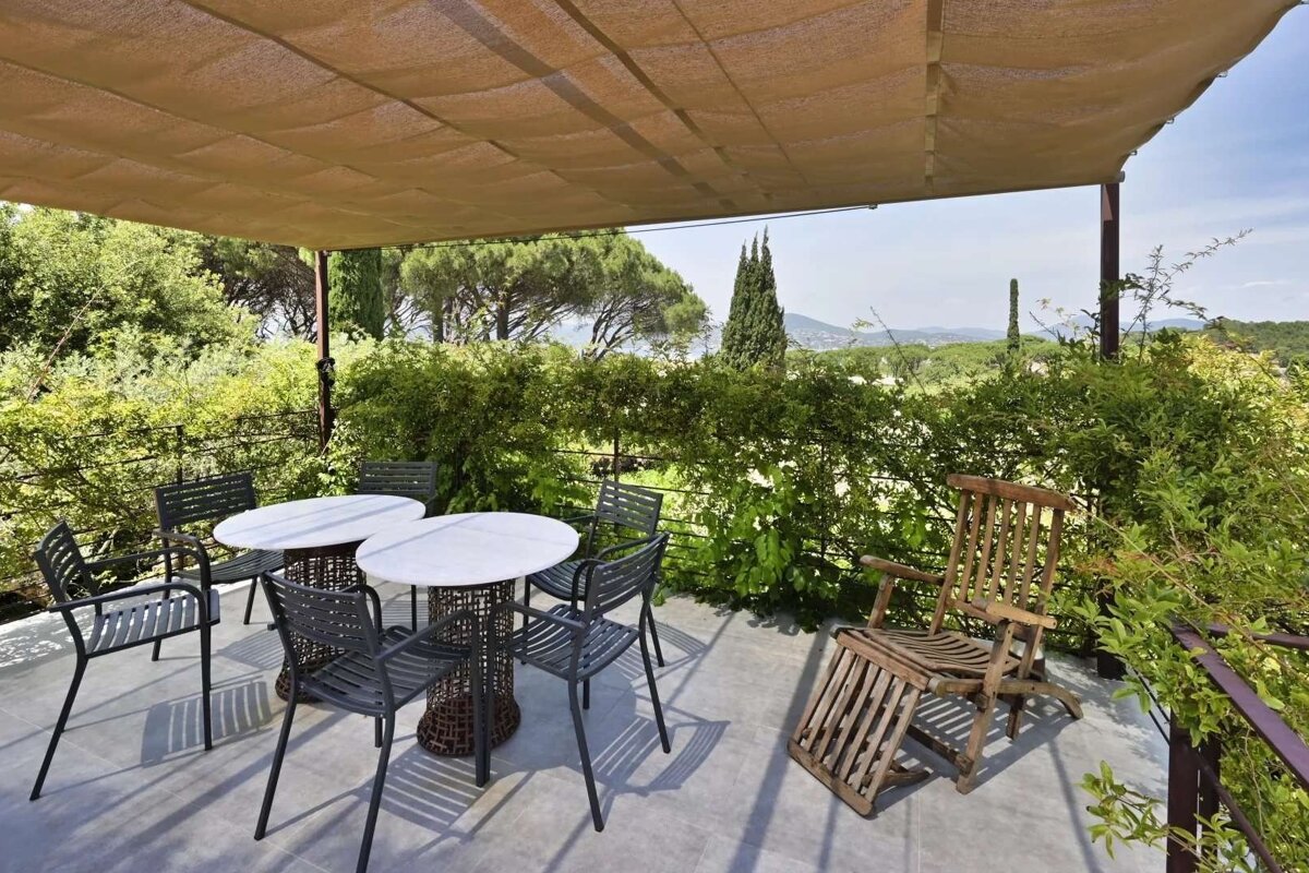 A shaded outdoor patio with two tables, chairs, and a wooden recliner, overlooking a lush, green landscape under a bright sky.