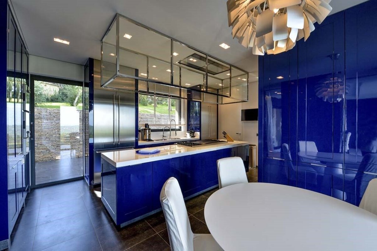 Vibrant modern kitchen featuring glossy royal blue cabinets, stainless steel appliances, a sleek dining area, and large windows with outdoor views.