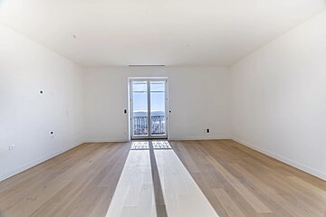 An empty white room with light wood floors and sunlight streaming in through a central glass balcony door.