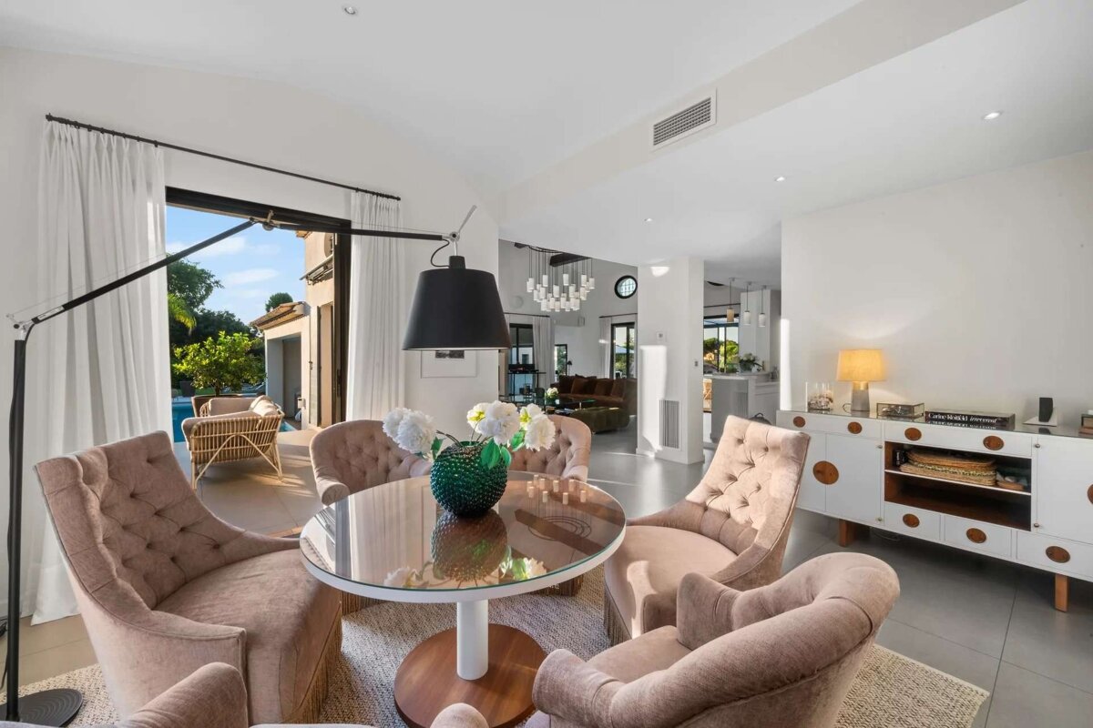 Bright, open-plan living area featuring a dining space with tufted chairs, a glass table, and a scenic view of an outdoor pool.