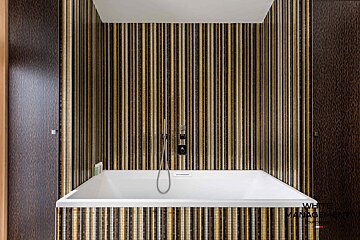 A white rectangular bathtub in an alcove with gold, brown, and black vertically striped mosaic tile walls.