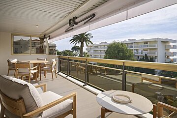 A sunny balcony with wooden furniture, dining table, and glass railing overlooking apartment buildings and palm trees.