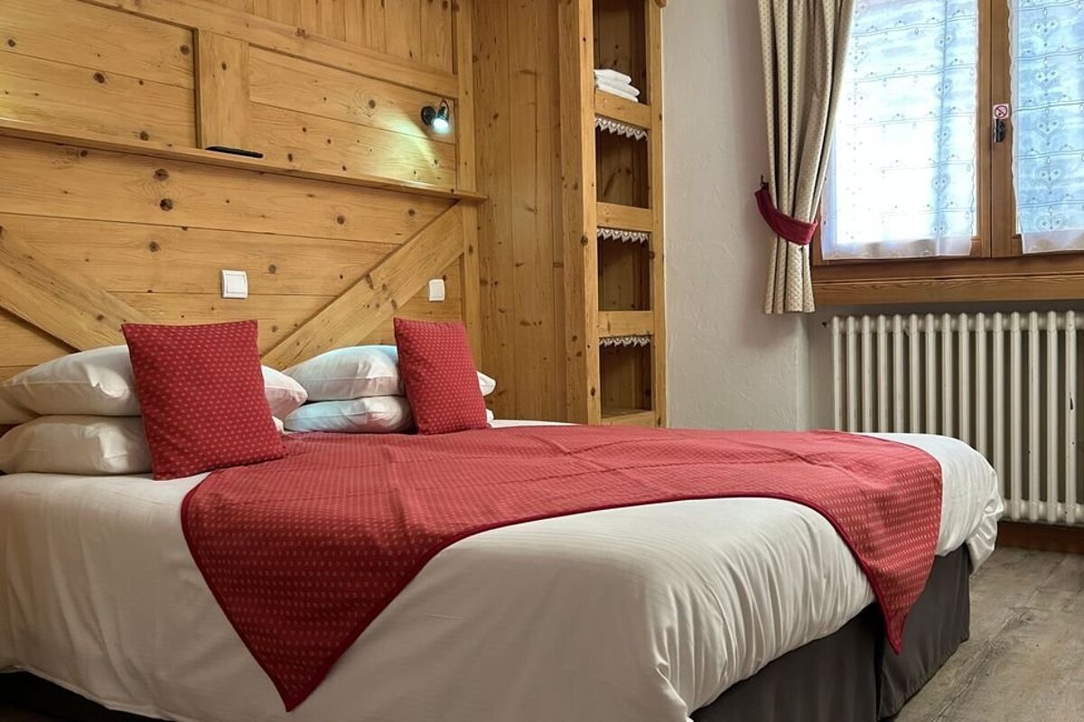Rustic bedroom with wood-paneled walls, featuring a bed made with white linens and red pillows and throw next to a window.