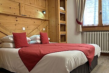 Rustic bedroom with wood-paneled walls, featuring a bed made with white linens and red pillows and throw next to a window.
