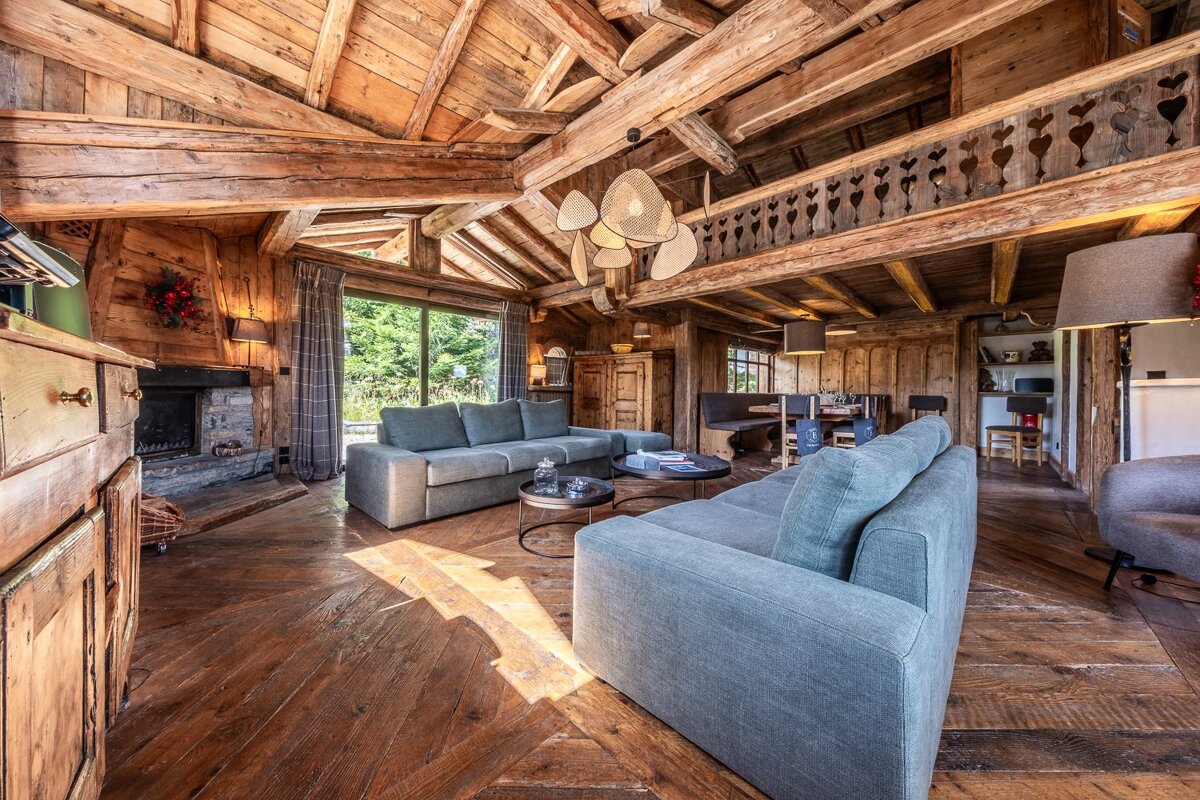 A warm, rustic chalet living room with exposed wooden beams, grey sofas, a stone fireplace, and large windows framing a lush forest view.
